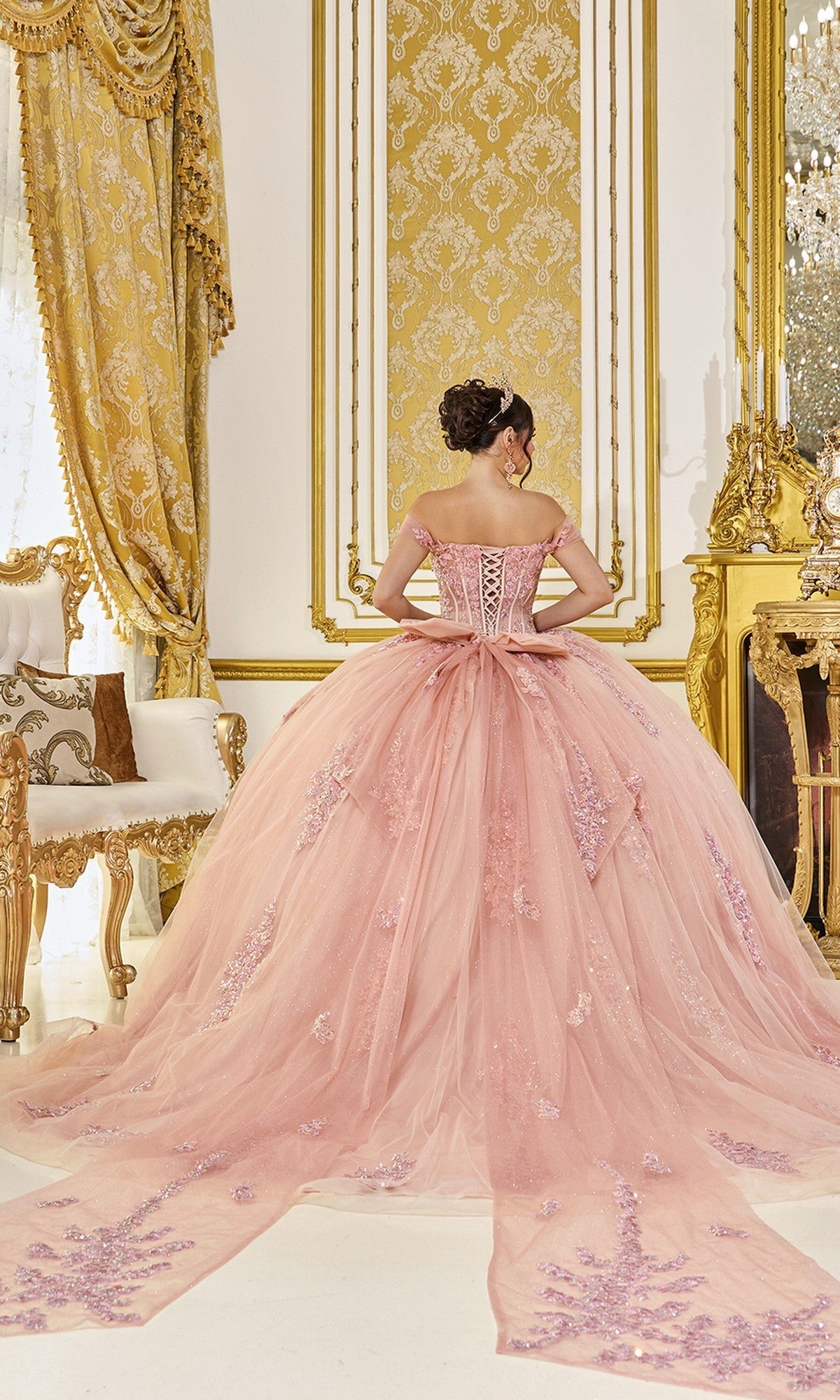 A woman in the Quinceanera Dress 5121 By Dancing Queen, featuring ornate rose gold floral lace and an off-the-shoulder neckline, stands with her back to the camera in a lavish, gold-accented room with elaborate decor.